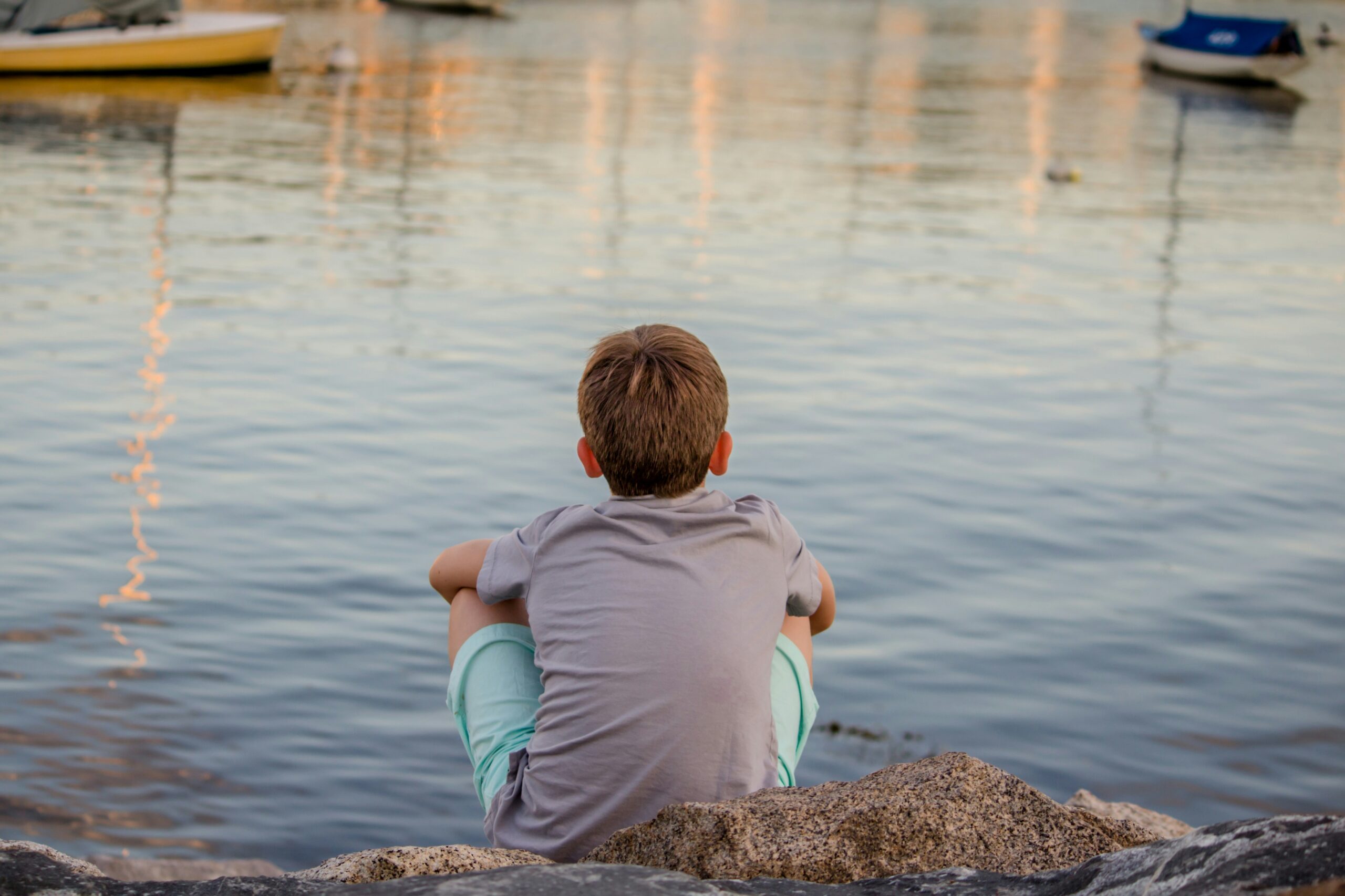 a young boy sits on a breakwater overlooking boats in a marina